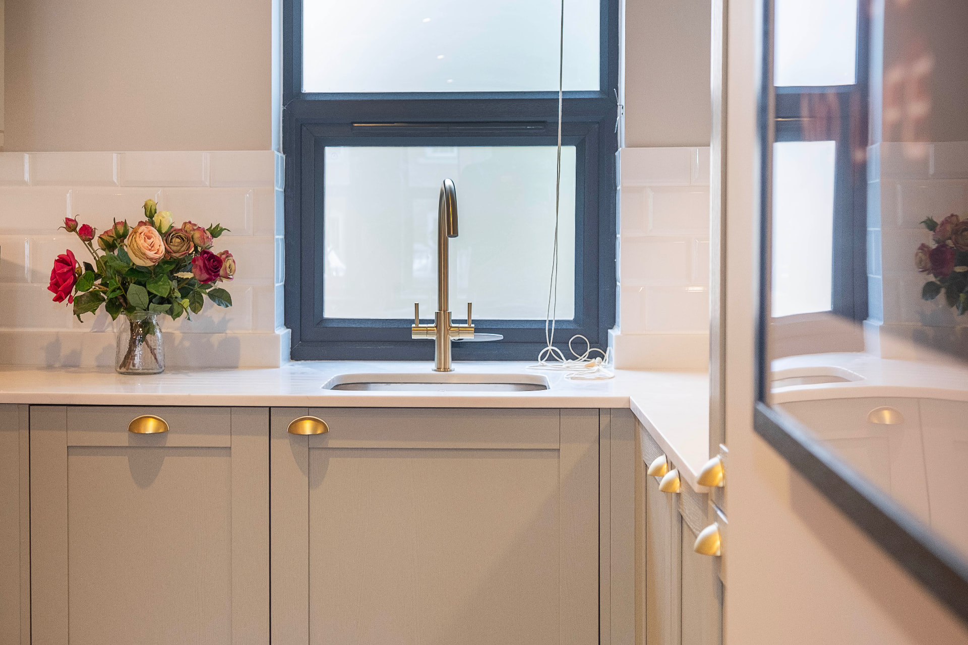 Brass kitchen tap and sink beneath a dark framed window, with flowers and a tiled splashback